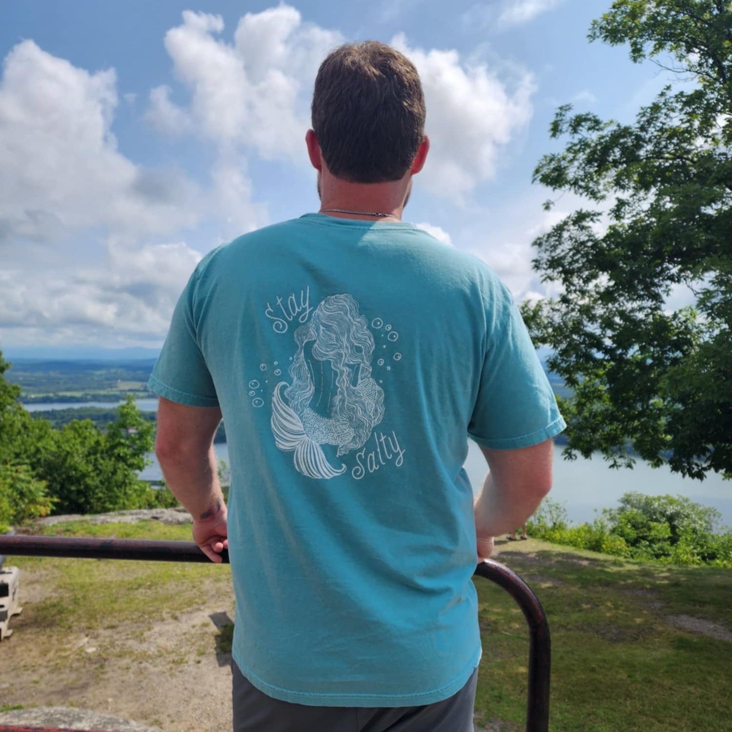 Person wearing a turquoise t-shirt with a mermaid design, standing on a scenic overlook.